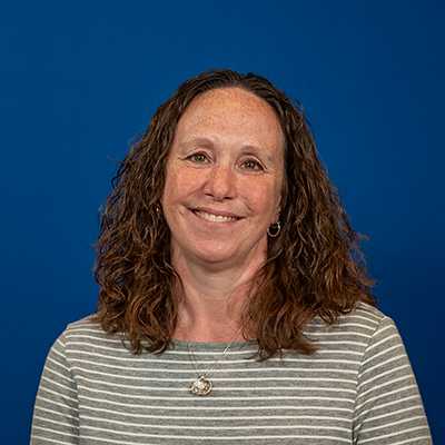 A white woman with curly brown hair, smiling, wearing a grey and white striped shirt.