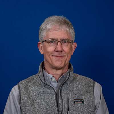 An older white man with glasses and short white hair wearing a grey shirt and vest.