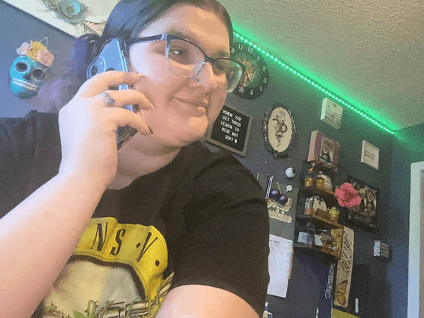 Young woman wearing glasses and a black T-shirt talking on a blue smartphone in a room decorated with dark walls and various wall hangings.