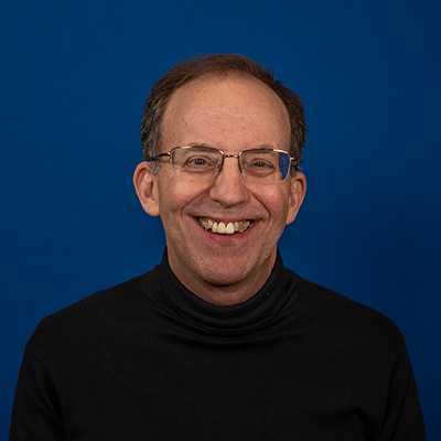 A white man in his 60s with thinning brown hair and glasses, wearing a black turtleneck shirt, smiling at the camera.
