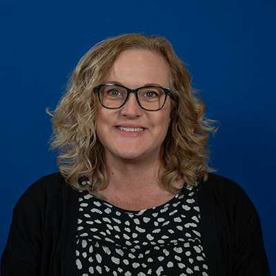 A white woman in her 40s with blond wavy hair who is wearing glasses and smiling.