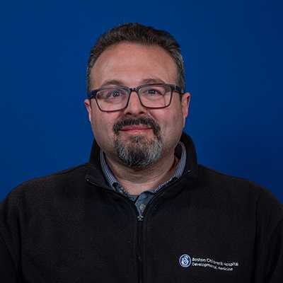 A middle-aged white male with salt-and-pepper hair and beard wearing black Boston Children’s Hospital fleece over a dress shirt and glasses.