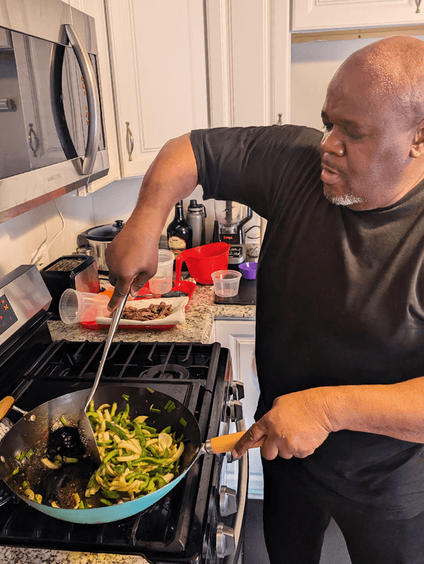 A bald Black man in a black shirt stirs vegetables in a pan on a stovetop in the kitchen.